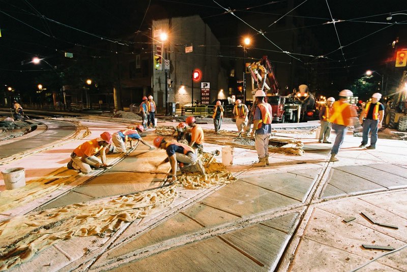 Various construction workers finishing concrete on streetcar tracks at night.